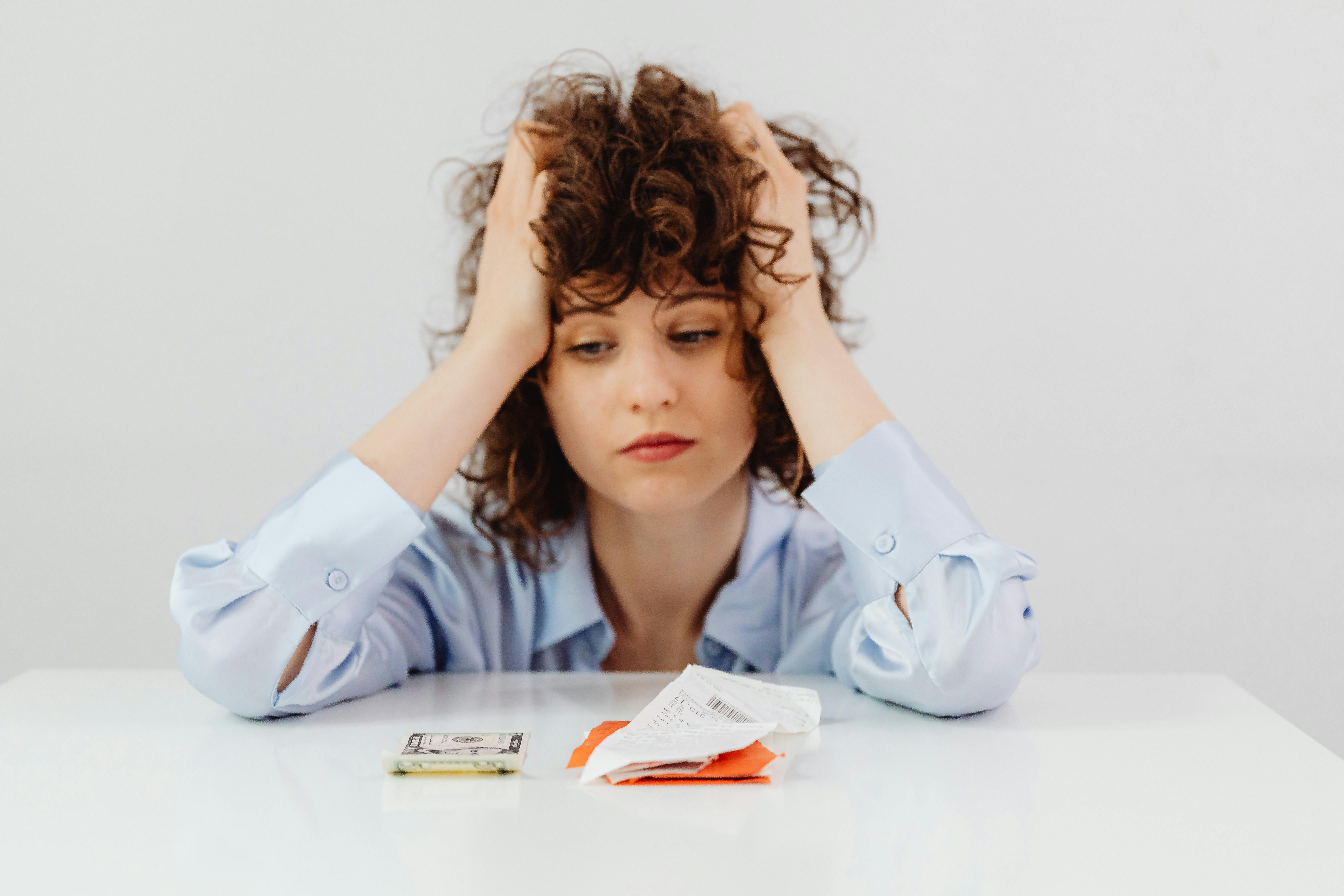 Exasperated woman leaning on a table with her hands in her hair, staring at a stack of receipts and a single dollar bill.